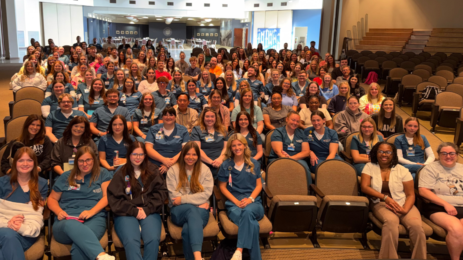 A large group of nursing students, many wearing matching teal scrubs, seated together in a tiered auditorium for a class photo.